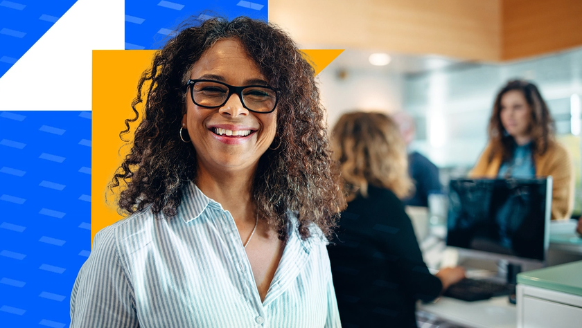 A smiling woman with glasses stands confidently in an office environment, with coworkers in the background.