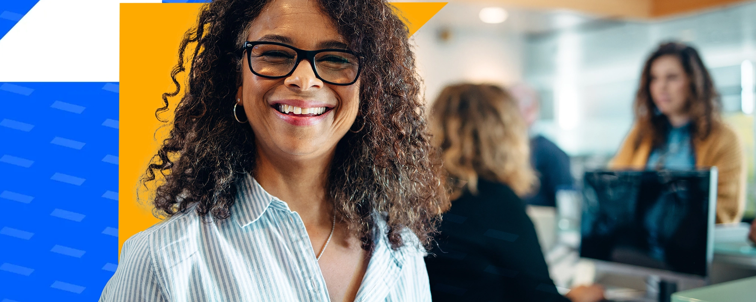 A smiling woman with glasses stands confidently in an office environment, with coworkers in the background.